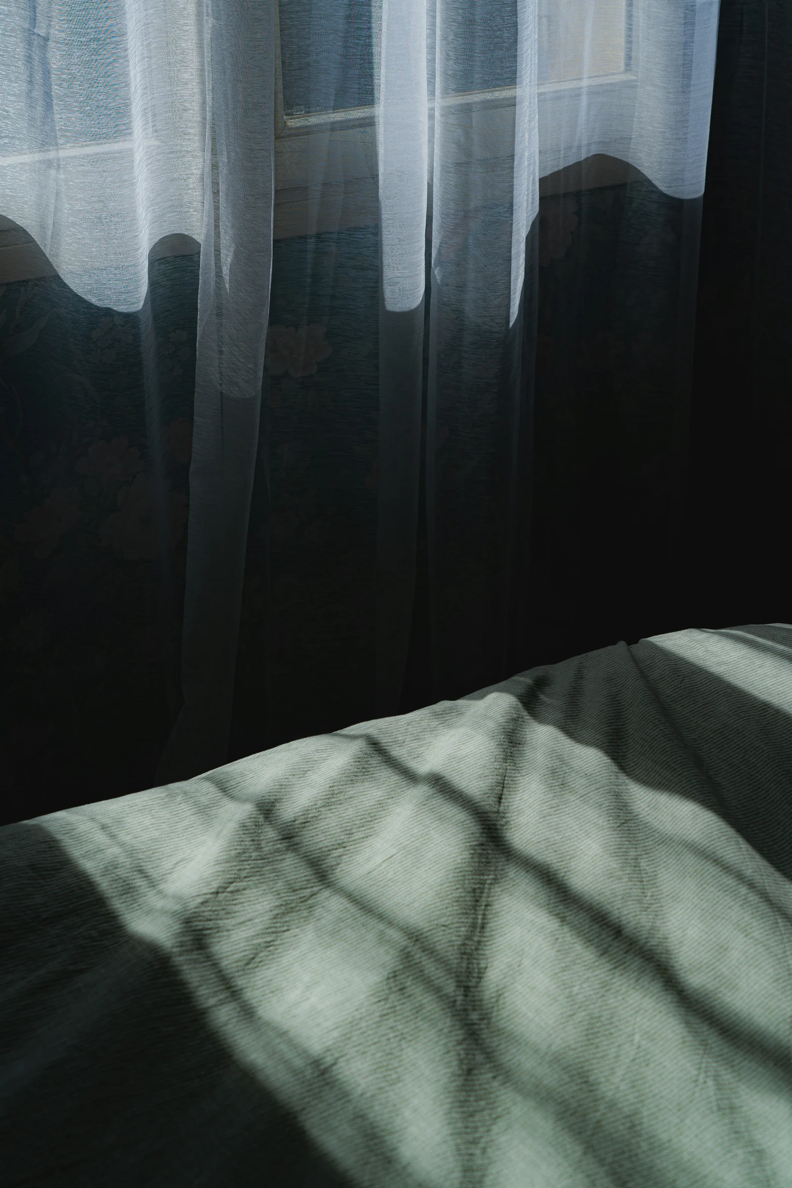Soft window light on curtains and bed linen in a dark bedroom.