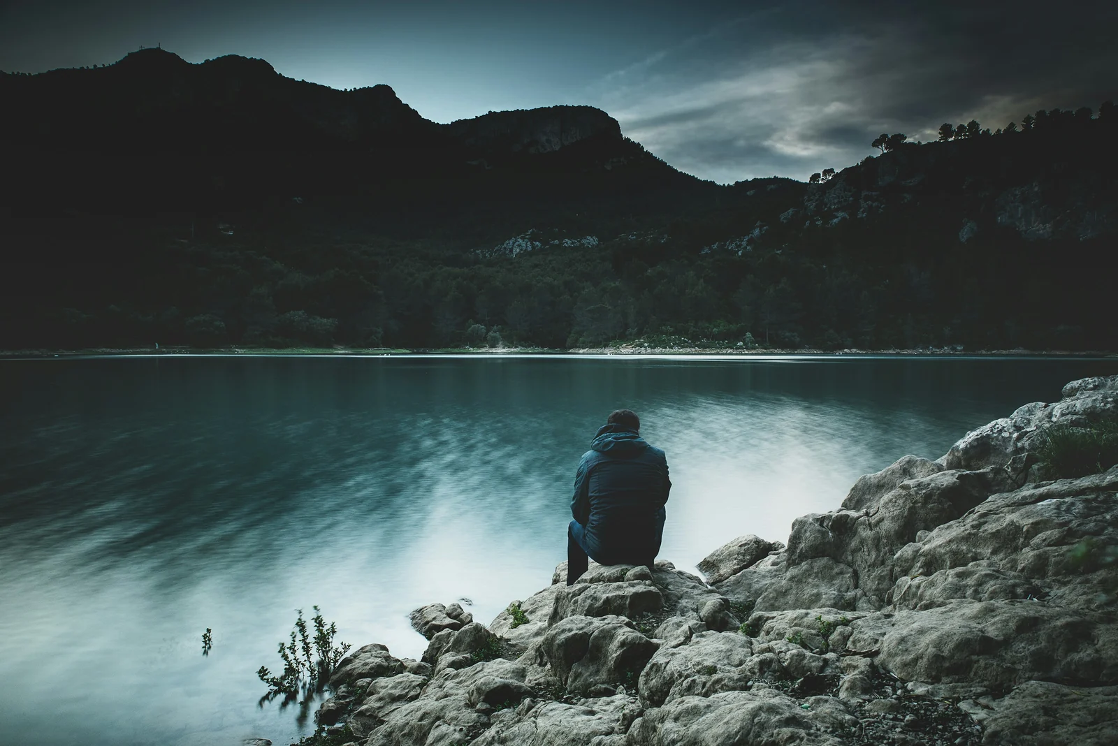 A person sits facing a dark mountain lake with their back to the camera.